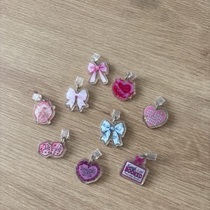 Op de afbeelding: Een verzameling van negen doorzichtige acryl bedels met diverse ontwerpen, waaronder roze en blauwe strikken, een roze hart met tekst, een roze dobbelsteenpaar, een skelet en een rechthoekig bordje met de tekst Sorry I Am BOOKED Please Leave Me Alone.