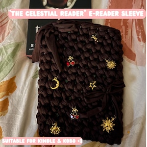 Op de afbeelding: Een donkerbruine gehaakte e-reader hoes met een lint en diverse goudkleurige bedels waaronder zonnen, sterren, een maansikkel en rode hartvormige kersen. De tekst luidt THE CELESTIAL READER E-READER SLEEVE en SUITABLE FOR KINDLE & KOBO.