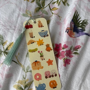 May include: A light beige bookmark featuring food-themed illustrations such as cherries, a croissant, and a donut. The bookmark has a light green tassel and a small red cherry charm. The background is a floral pattern.