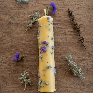May include: A cylindrical yellow beeswax candle embedded with small purple dried flowers and green sprigs. The candle features a single central wick at the top and is surrounded by loose, scattered dried purple flower heads and green foliage.