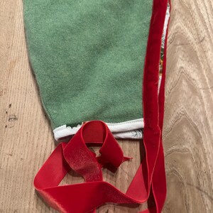 May include: A green baby bonnet with a red velvet trim and matching ribbon. The bonnet is made of a soft, textured fabric and has a white trim along the edge. The red ribbon is curled and lies on a wooden surface.
