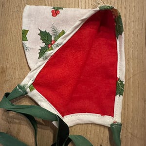 Puede incluir: Un gorro navideño festivo con interior rojo y exterior blanco estampado con hojas y bayas de acebo. El gorro está ribeteado con tela blanca y tiene lazos verdes.