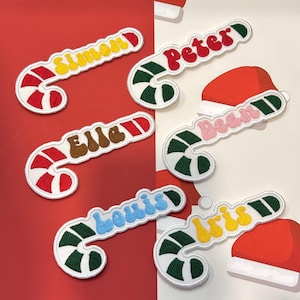 May include: Embroidered candy cane patches with names in various colors. The candy canes have red and white stripes, green and white stripes, and white borders. Names include Simon, Peter, Elle, Dean, Louis, and Iris.