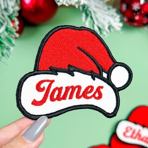 May include: A red and white embroidered patch shaped like a Santa hat with a black border. The white brim of the hat features the name James stitched in red cursive script. The patch has a soft texture and a distinct, bold outline design.