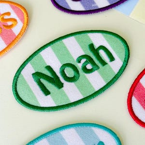 May include: An oval-shaped embroidered patch featuring a white and light green vertical striped pattern. The name Noah is stitched in dark green thread across the center of the patch, which is finished with a dark green stitched border edge.