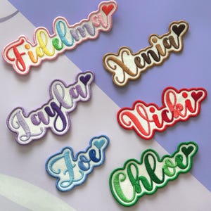May include: A selection of embroidered name patches in a range of colours, such as rainbow, purple, red, and green. Each patch displays a different name in a cursive font, with a heart detail. Names include Fidelma, Xenia, Layla, Vicki, Joe, and Chloe.