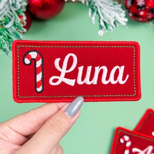 May include: A rectangular red embroidered patch featuring a white candy cane graphic on the left side and the name Luna in white cursive script lettering. The patch has a green stitched border and a red fabric background with a textured finish.
