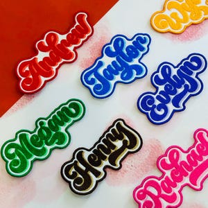 May include: A collection of embroidered name patches in various colors, including red, blue, green, brown, pink, and yellow. Each patch features a different name in a cursive font with a white border. The patches are arranged on a white and red background.