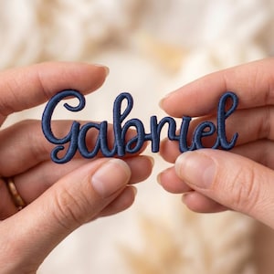 May include: A navy blue embroidered patch with the name "Gabriel" in a cursive script. The patch is held between two hands, showcasing the detailed stitching and color of the lettering. The background is a soft, blurred neutral tone.