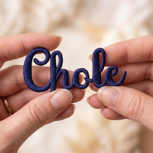 May include: A dark navy blue embroidered patch featuring the word "Chole" in a cursive, connected script font. The lettering is thick and textured with visible thread stitching, creating a raised appearance for the custom name design.