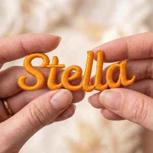 May include: A bright orange embroidered patch featuring the name Stella in a cursive script font. The letters are connected in a single piece with a raised, textured thread finish, showcasing a vibrant color and detailed needlework throughout.