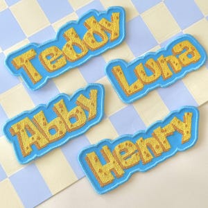 May include: Four embroidered name patches: "Teddy", "Luna", "Abby", and "Henry". Each patch features yellow text with a cheese-like texture, outlined in brown, set against a light blue background. The patches are on a checkered surface.