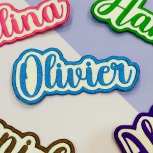 May include: Embroidered name patches in various colors. The patch in focus says "Olivier" in blue script with a white background and a blue border. Other patches with names are visible in pink, green, brown, and purple.