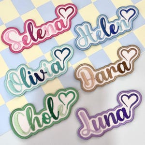 May include: Embroidered name patches in various colors, including pink, blue, green, brown, and purple. Each patch features a name in a cursive font with a heart-shaped accent. The patches are displayed on a checkered background.
