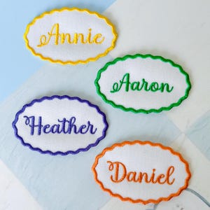May include: Four embroidered name patches with white ovals and scalloped edges. Each patch features a different name in a cursive font: "Annie" in yellow, "Aaron" in green, "Heather" in purple, and "Daniel" in orange. The patches are on a light blue and white patterned surface.