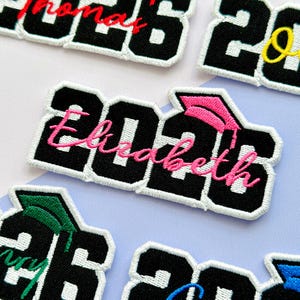 May include: Embroidered patches with the year "2026" in black block letters with a white border. The name "Elizabeth" is stitched in pink cursive. A pink graduation cap is above the "2026". Other patches are visible.