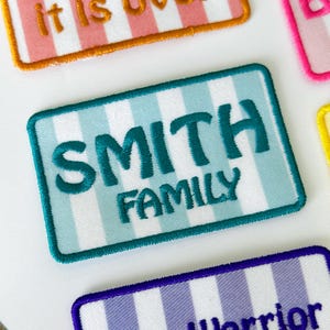 May include: A rectangular embroidered patch featuring a light blue and white vertical striped background with a dark teal border. The text SMITH FAMILY is embroidered in dark teal capital letters across the centre of the patch.