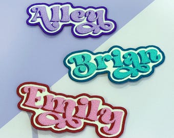 May include: Three embroidered name patches in a retro font. The names are "Alley" in purple, "Brian" in teal, and "Emily" in pink. Each patch has a contrasting border and is set against a lavender and white background.
