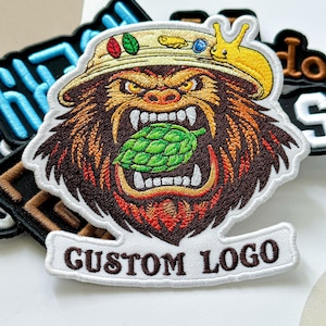 May include: Embroidered patch featuring a fierce gorilla wearing a fishing hat and biting a green hop. The patch has a white border with the words "CUSTOM LOGO". The gorilla is brown with orange and yellow accents.
