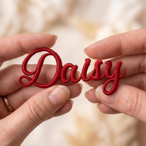 May include: A red embroidered patch with the name "Daisy" in a cursive font. The patch is held between two hands against a soft, blurred background. The letters are connected and have a slight sheen.