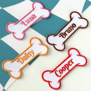 May include: Four embroidered dog bone patches with names: Luna, Bruno, Daisy, and Cooper. Each bone is white with a colored border and matching text. The patches are arranged on a blue and white checkered surface.