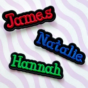 May include: Three embroidered name patches: "James" in red, "Natalie" in blue, and "Hannah" in green. Each name is stitched in a bold, cursive font with a black border. The patches are on a striped background.