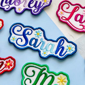 May include: Embroidered name patches in various colours. The name "Sarah" is in blue, with a white background and blue outline. Small flower details are scattered around the name. Other names include "Lorey" and "La" in different colours and fonts.