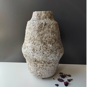Könnte beinhalten: Eine strukturierte, beige Vase mit breitem Boden und schmalem Hals. Die Vase hat ein raues, organisches Aussehen mit sichtbarer Textur auf ihrer Oberfläche. Die Vase wird auf einer weißen Oberfläche vor einem dunkelgrauen Hintergrund ausgestellt.