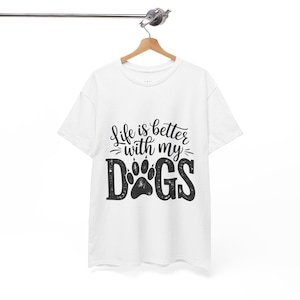 Può includere: Una t-shirt bianca a maniche corte con la scritta nera Life is better with my DOGS, in cui la lettera O è sostituita da un'impronta di zampa nera. Il capo è appeso a una gruccia di legno su uno sfondo bianco uniforme.