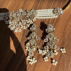 May include: A gold-toned jewelry set featuring a choker necklace and matching earrings. The necklace has multiple strands of white pearls and a central ornate design with clear gemstones. The earrings mirror the design, with dangling pearl accents.