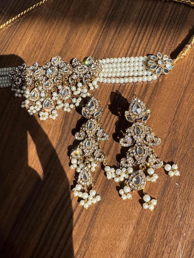 May include: A gold-toned jewelry set featuring a choker necklace and matching earrings. The necklace has multiple strands of white pearls and a central ornate design with clear gemstones. The earrings mirror the design, with dangling pearl accents.
