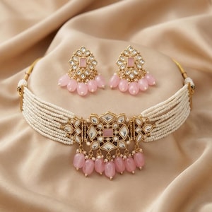 Könnte beinhalten: Ein goldfarbenes Halsketten- und Ohrring-Set mit einem mehrsträngigen, weiß perlenbesetzten Choker. Das Set enthält passende Ohrringe und einen Anhänger mit rosa Edelsteinakzenten und einem floralen Design. Der Schmuck wird auf einer beigen Satin-Oberfläche präsentiert.