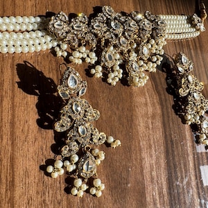 Pode incluir: Um conjunto de joias ornamentado com um colar gargantilha de pérolas, brincos a condizer e um pendente. As peças de metal dourado são adornadas com cristais transparentes e pequenas pérolas penduradas, criando um visual luxuoso e elegante.