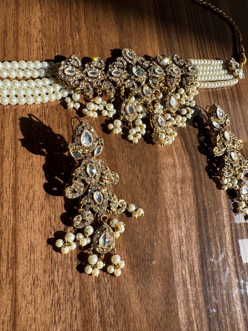 May include: An ornate jewelry set featuring a pearl choker necklace, matching earrings, and a pendant. The gold-toned metal pieces are adorned with clear crystals and small, dangling pearls, creating a luxurious and elegant look.