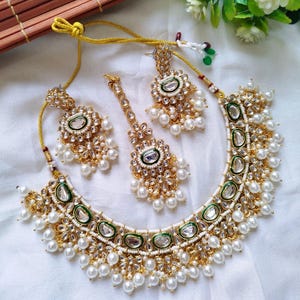 Puede incluir: Un conjunto de joyas dorado que incluye un collar, pendientes y una pieza para la cabeza. El conjunto está adornado con perlas blancas, piedras transparentes y detalles verdes. El collar y los pendientes tienen un diseño floral.