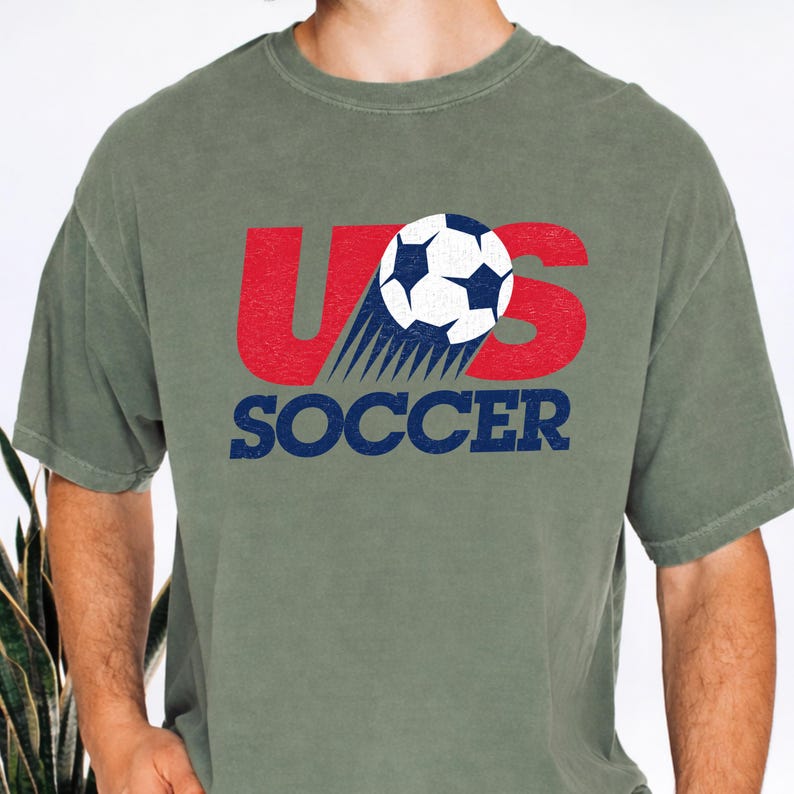Op de afbeelding: Een olijfgroen T-shirt met korte mouwen en een grafische print met de rode letters US, een wit-blauwe voetbal die de letter S vervangt, en het woord SOCCER in blauwe tekst eronder. Het ontwerp heeft een verweerde, vintage textuur.