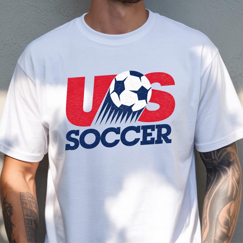 Op de afbeelding: Een wit T-shirt met korte mouwen en een grafisch ontwerp op de borst. Het ontwerp bevat de rode letters U en S, waarbij een zwart-witte voetbal de letter S vervangt, gevolgd door het woord SOCCER in dikke blauwe hoofdletters.
