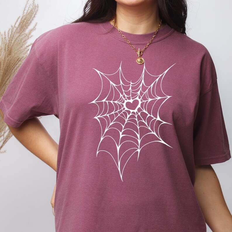 May include: A maroon t-shirt featuring a white spiderweb design with a heart in the center. The shirt has short sleeves and a crew neck. The fabric appears soft and comfortable.