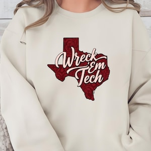 May include: Cream-colored sweatshirt featuring a maroon Texas-shaped graphic with a floral pattern. The graphic includes the words "Wreck 'Em Tech" in a stylized font, a nod to Texas Tech University.