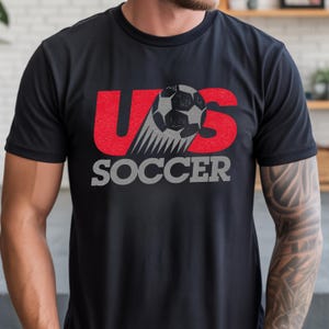Op de afbeelding: Een zwart t-shirt met korte mouwen en een grafisch ontwerp met de rode letters U en S. Een grijze en zwarte voetbal is tussen de letters geplaatst met bewegingslijnen, en het woord SOCCER staat in grijze blokletters eronder gedrukt.