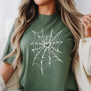 May include: A green t-shirt featuring a white spiderweb design with a heart-shaped center. The shirt has short sleeves and a crew neck. The design is centered on the front of the shirt.