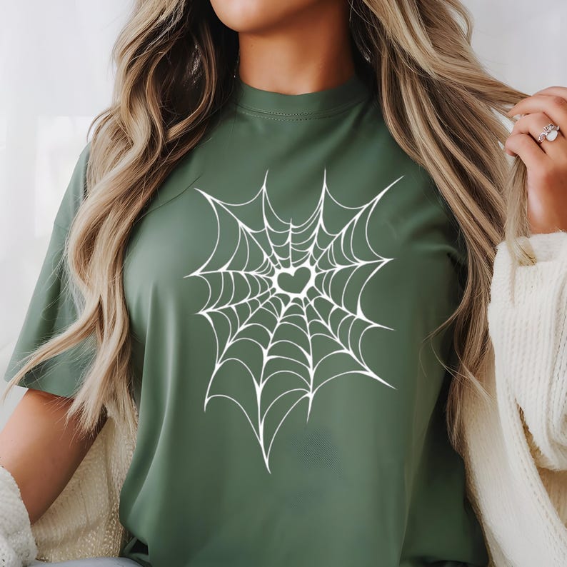 May include: A green t-shirt featuring a white spiderweb design with a heart-shaped center. The shirt has short sleeves and a crew neck. The design is centered on the front of the shirt.