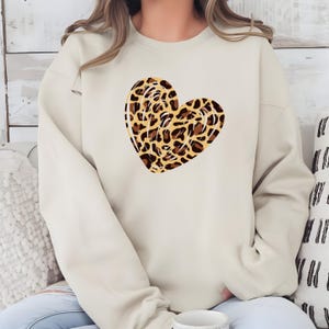 May include: A cream-colored sweatshirt featuring a large heart design. The heart is filled with a leopard print pattern in shades of brown and tan. The sweatshirt has a crew neck and long sleeves.