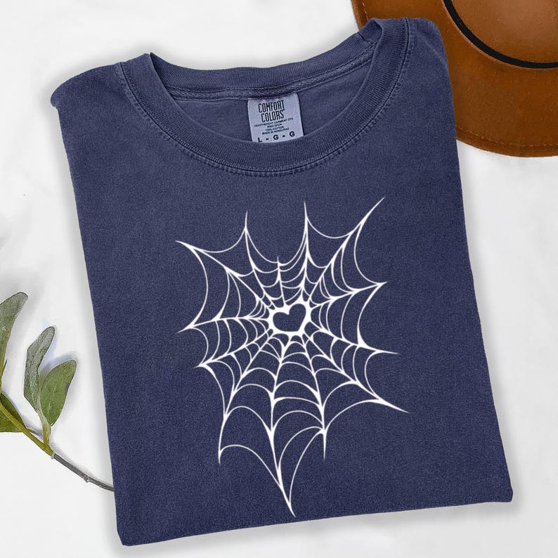 May include: A navy blue t-shirt with a white spiderweb design featuring a heart in the center. The shirt is folded and has a crew neck. The brand label "Comfort Colors" is visible.