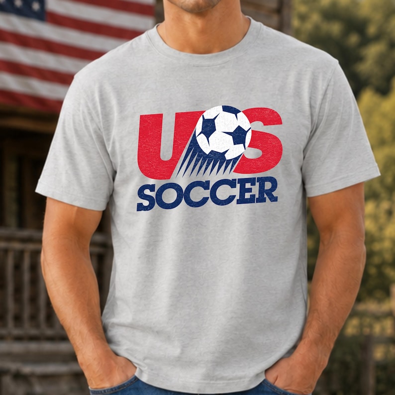 Op de afbeelding: Een lichtgrijs t-shirt met korte mouwen en een grafisch ontwerp met de tekst US SOCCER in rood en blauw. Een zwart-witte voetbal is boven het woord SOCCER geplaatst met bewegingslijnen erachter, wat een dynamisch effect cre&euml;ert.
