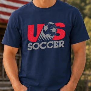 Op de afbeelding: Een marineblauw t-shirt met korte mouwen en een grafische print met de rode letters U en S, een grijze voetbal die de letter O vervangt, en het woord SOCCER in grijze blokletters eronder. De afbeelding heeft een verweerde textuur.