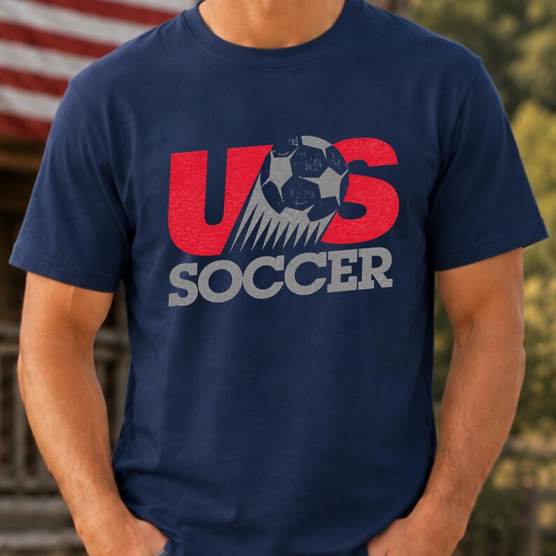 Op de afbeelding: Een marineblauw t-shirt met korte mouwen en een grafische print met de rode letters U en S, een grijze voetbal die de letter O vervangt, en het woord SOCCER in grijze blokletters eronder. De afbeelding heeft een verweerde textuur.