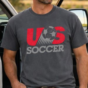Op de afbeelding: Een antracietgrijs t-shirt met korte mouwen en een grafisch ontwerp met de rode letters U en S. Een zwart-witte voetbal is tussen de letters geplaatst en het woord SOCCER staat eronder gedrukt in grote grijze hoofdletters.