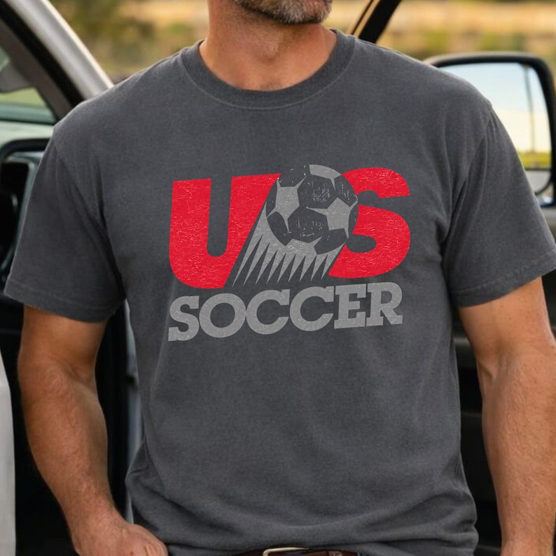 Op de afbeelding: Een antracietgrijs t-shirt met korte mouwen en een grafisch ontwerp met de rode letters U en S. Een zwart-witte voetbal is tussen de letters geplaatst en het woord SOCCER staat eronder gedrukt in grote grijze hoofdletters.