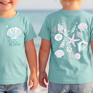 May include: A light teal t-shirt featuring a front graphic of a seashell with the name olivia and a back graphic displaying a collection of various seashells, a starfish, and seaweed in shades of pink, white, and teal against the fabric.